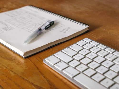 Image représentant une partie d'un clavier ainsi qu'un stylo sur un block note. Des caractères sont présents sur le bloc note. Le tout est posé sur un bureau en bois.